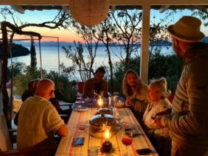 A group of friends and colleagues gather around a table on a terrace with a stunning view of the Peloponnese in the background.
