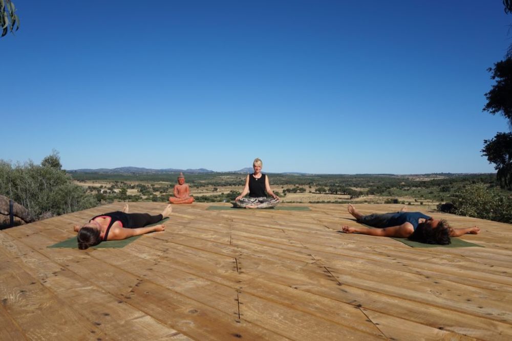 A yoga teacher sits in a cross-legged position on a wooden patio, teaching meditation to her students