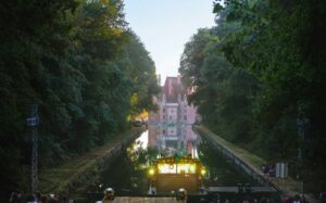 A music festival stage set up on a castle's lawn, surrounded by trees and a a tranquil pond in the foreground