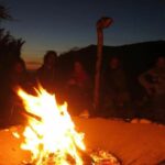 A group of people sit in a circle around a flickering campfire at night