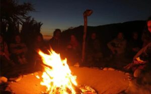 A group of people sit in a circle around a flickering campfire at night